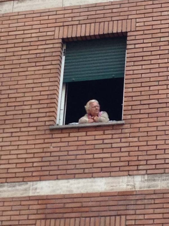 An old woman is looking out from her homes window of a brick building