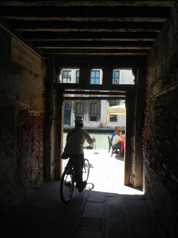 An old man biking outside a door in the ghetto area with the shadow behind him and the light infront of him at Venice Venezia 