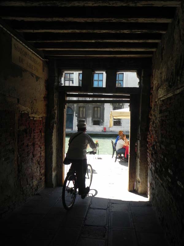 An old man biking outside a door in the ghetto area with the shadow behind him and the light infront of him at Venice Venezia